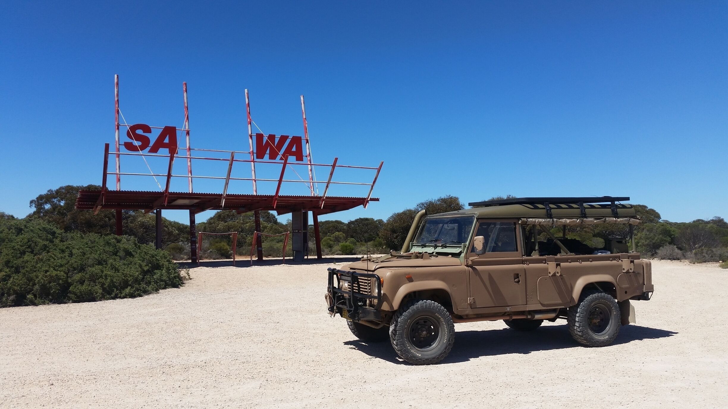A desert landscape with a tan off-road vehicle parked in front of a sign with 'SA' and 'WA' letters.