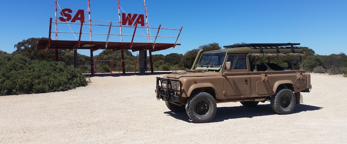 A desert landscape with a tan off-road vehicle parked in front of a sign with 'SA' and 'WA' letters.