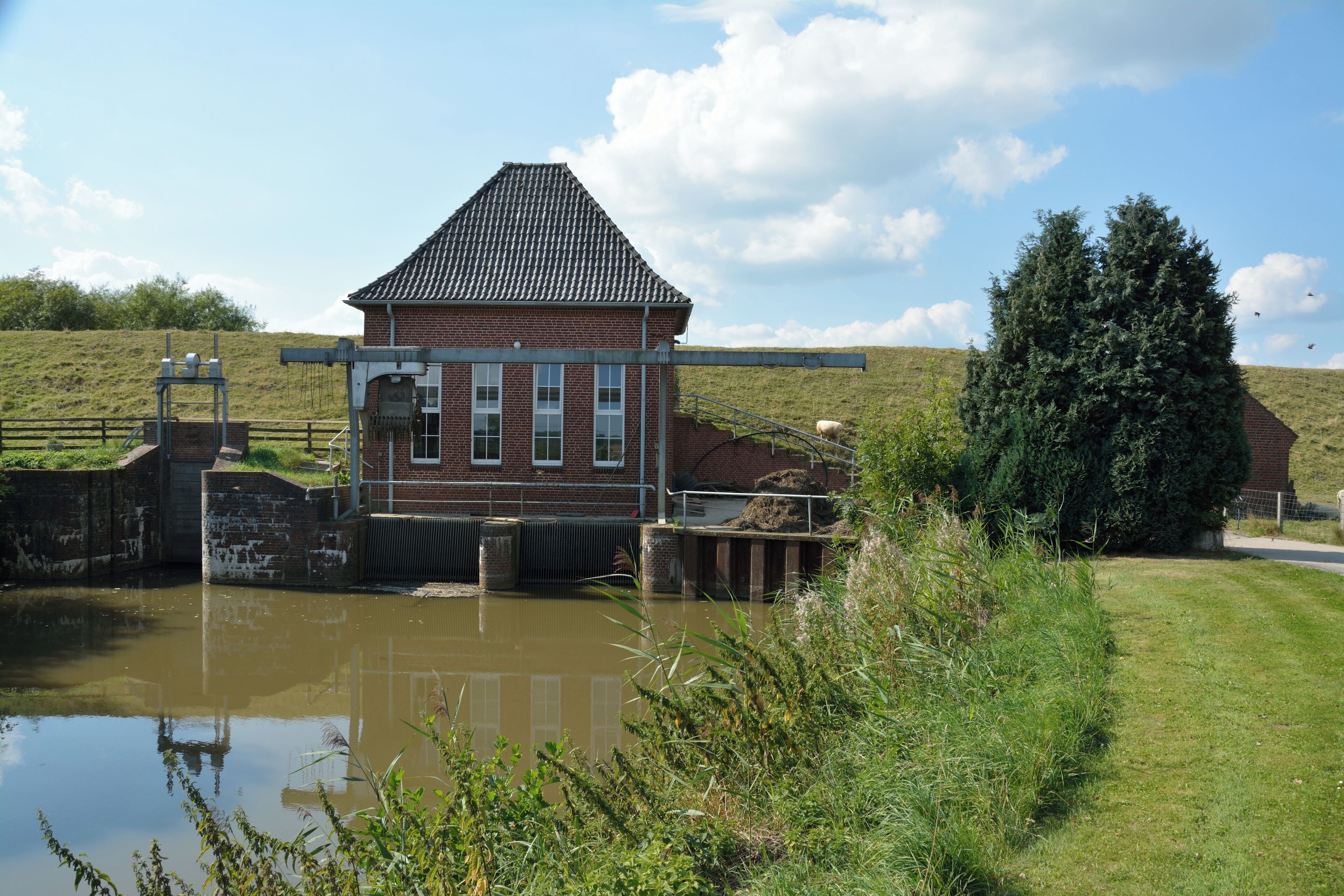A brick building with a tiled roof near a water body, surrounded by greenery and a clear sky.