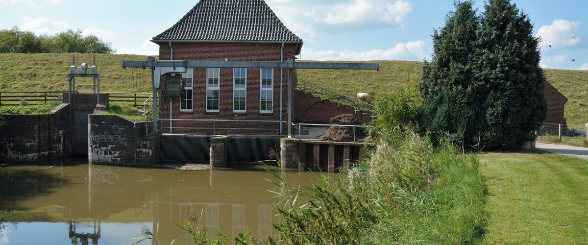 A brick building with a tiled roof near a water body, surrounded by greenery and a clear sky.