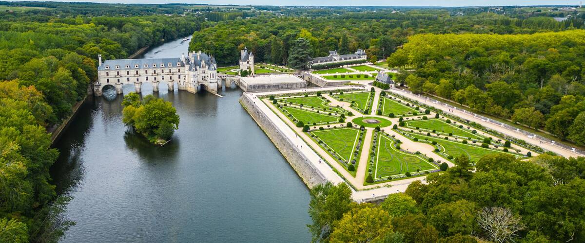 A historic castle with a river, formal gardens, and surrounding greenery.