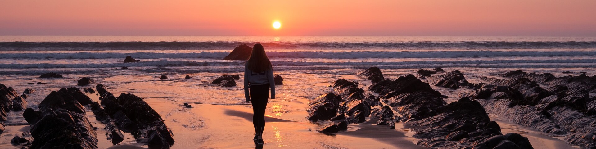 Un atardecer sobre una playa rocosa con olas y una persona de pie en la arena.
