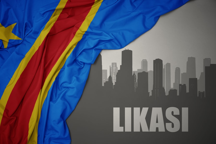 abstract silhouette of the city with text Likasi near waving colorful national flag of democratic republic of the congo on a gray background.