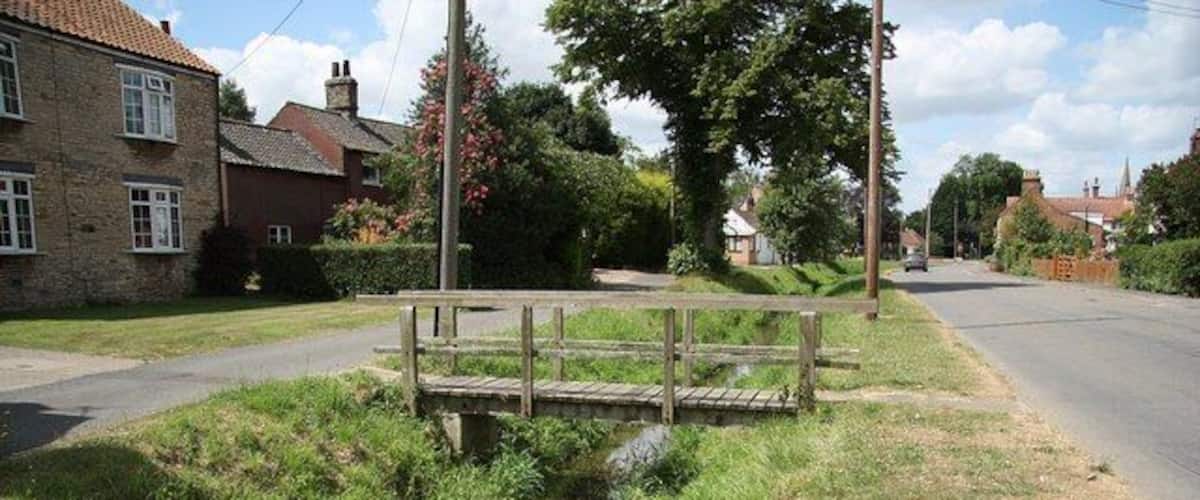 A rural road with a wooden bridge over a narrow stream, surrounded by houses and greenery.