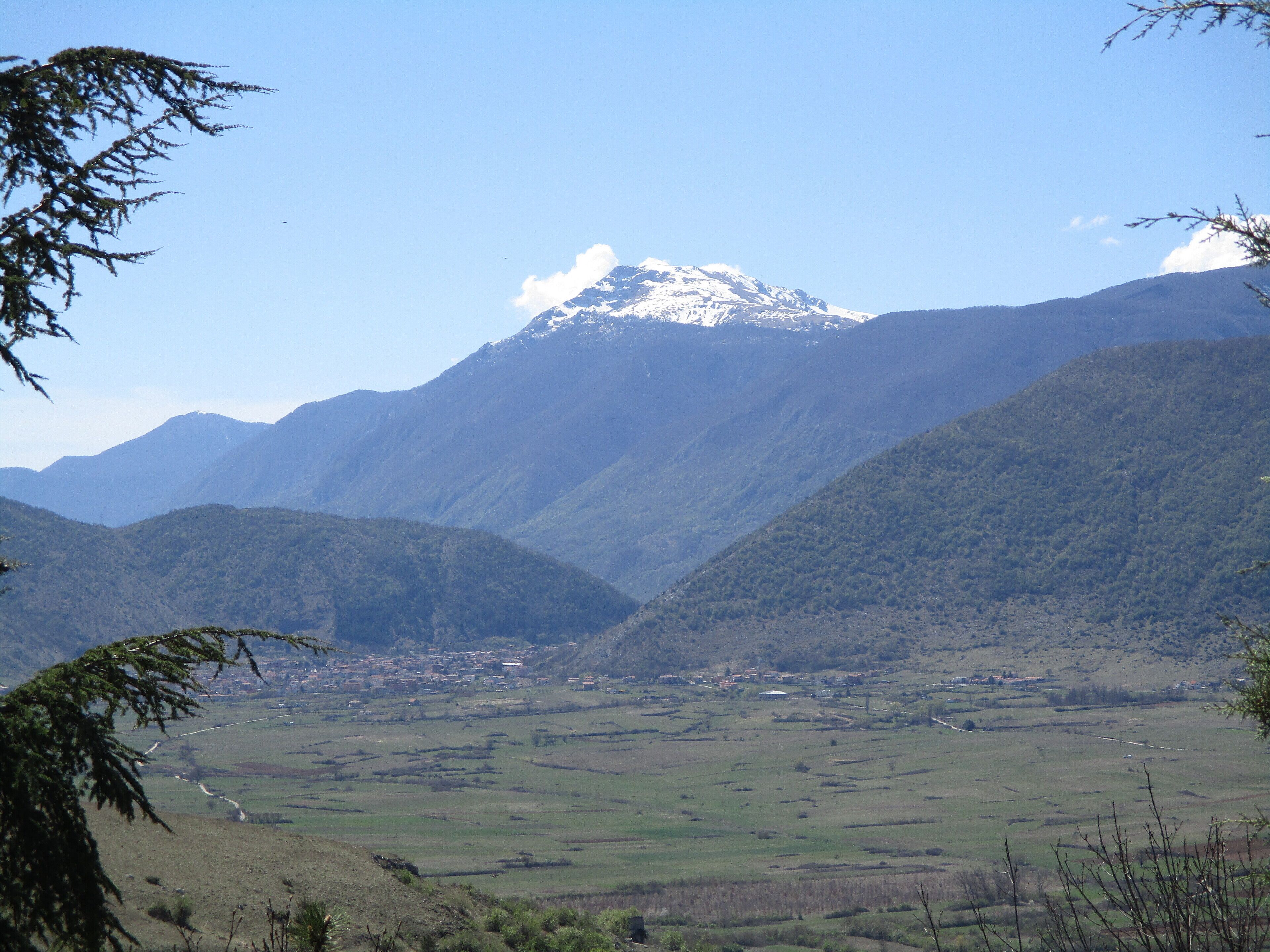 A mountainous landscape with a snow-capped peak, a valley with scattered buildings, and a clear blue sky.