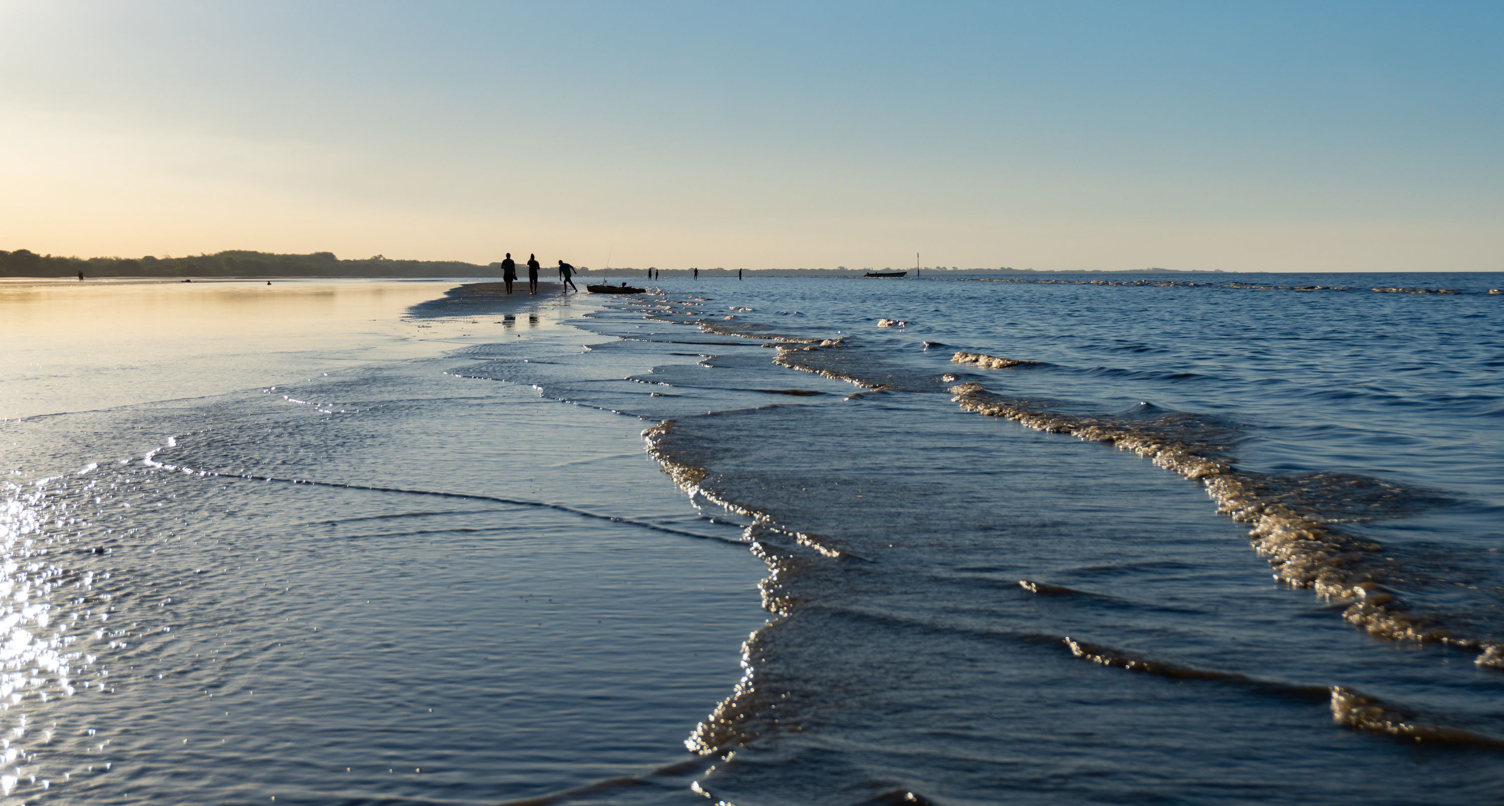 Una playa con olas suaves, cielo despejado y gente caminando a lo lejos.