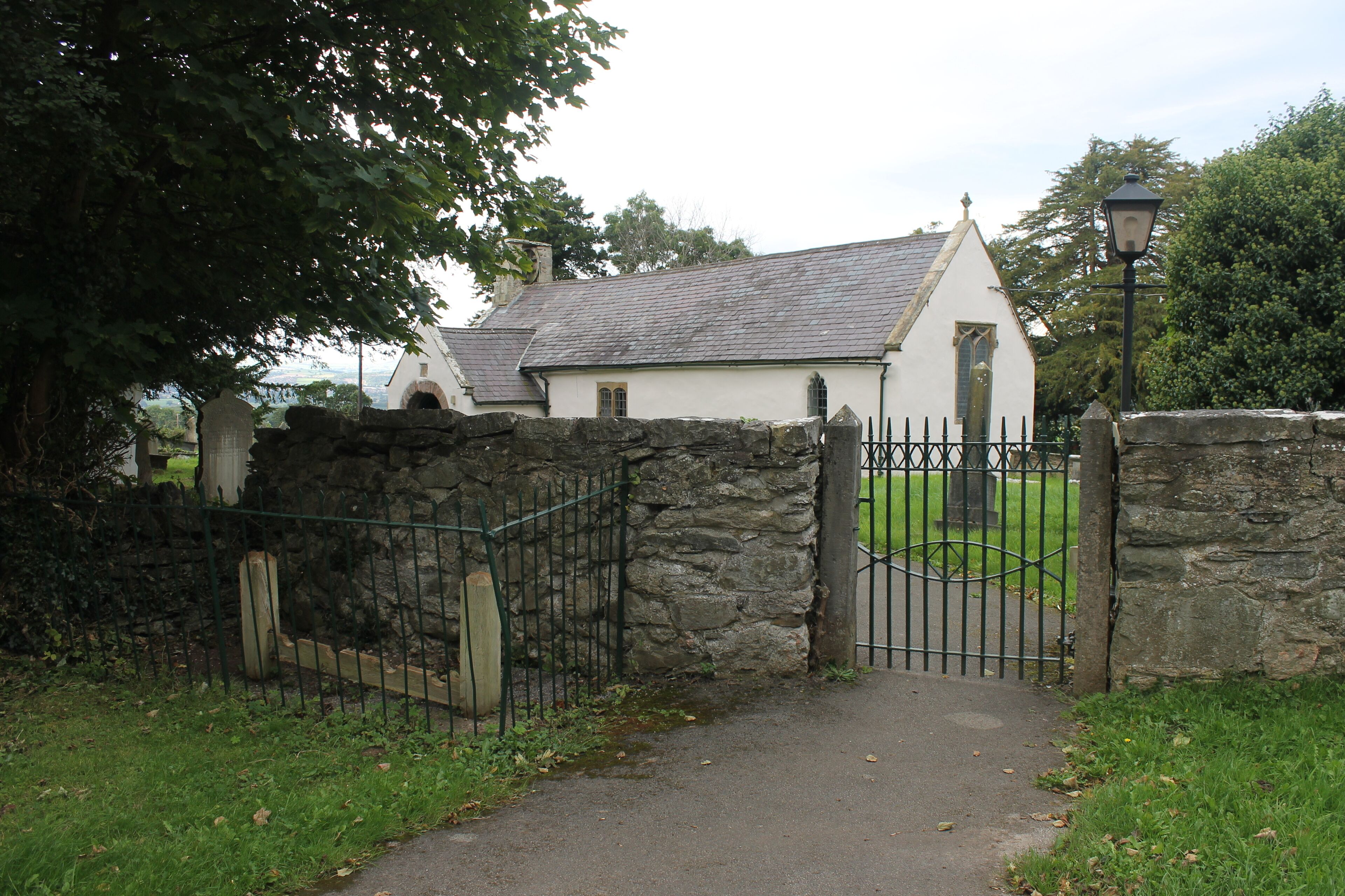 A stone gate leading to a white church with a grey roof, surrounded by greenery.