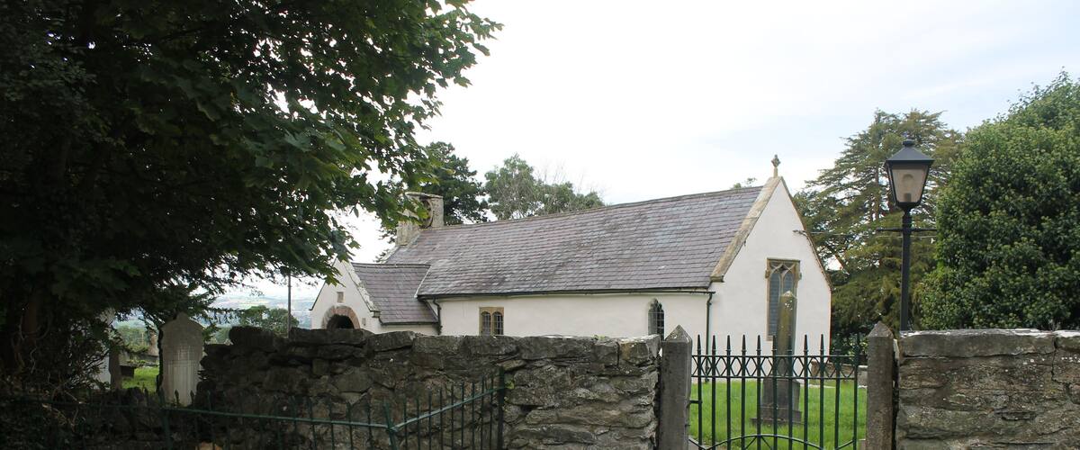 A stone gate leading to a white church with a grey roof, surrounded by greenery.