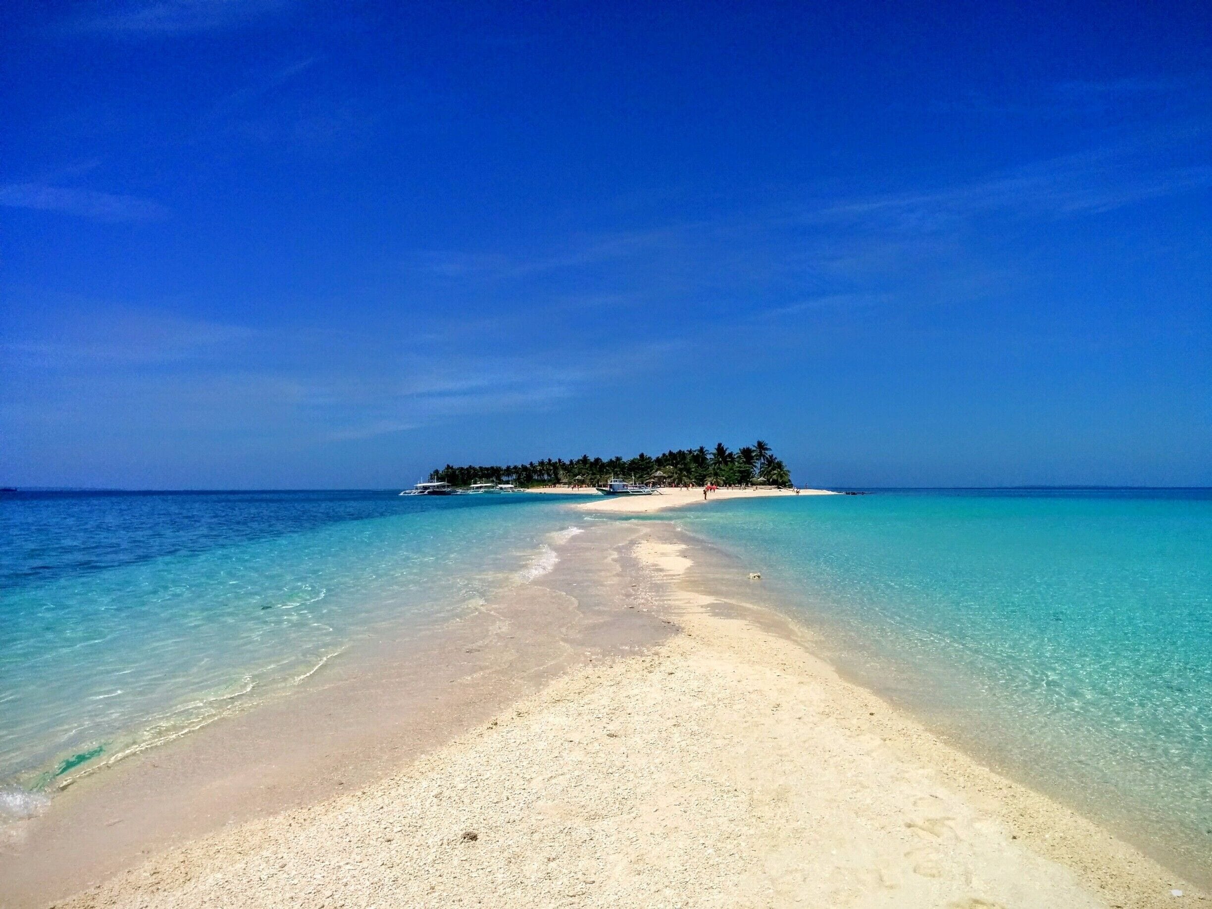 Una playa con aguas turquesas cristalinas, una orilla arenosa y una pequeña isla con palmeras al horizonte.