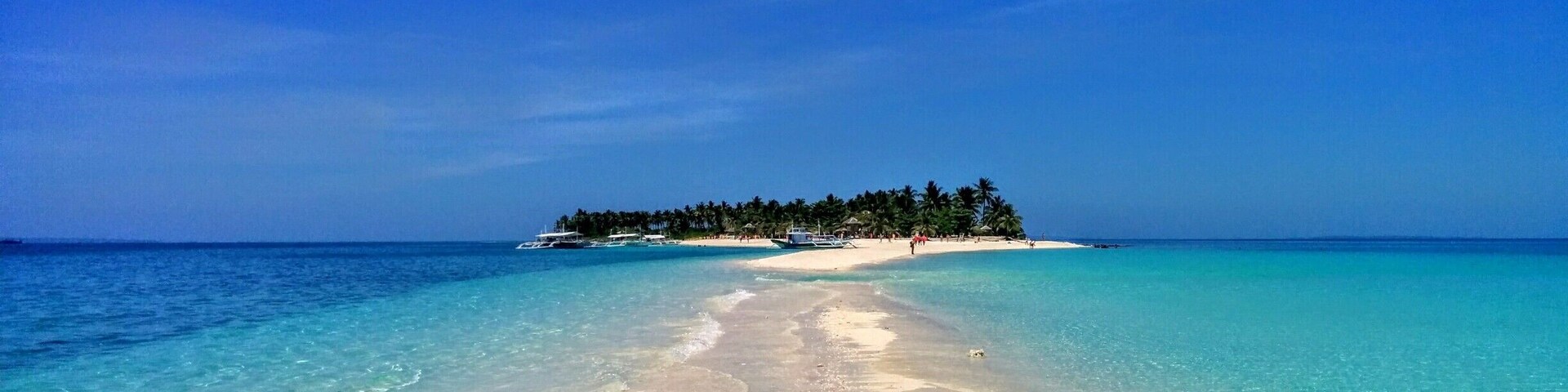Una playa con aguas turquesas cristalinas, una orilla arenosa y una pequeña isla con palmeras al horizonte.