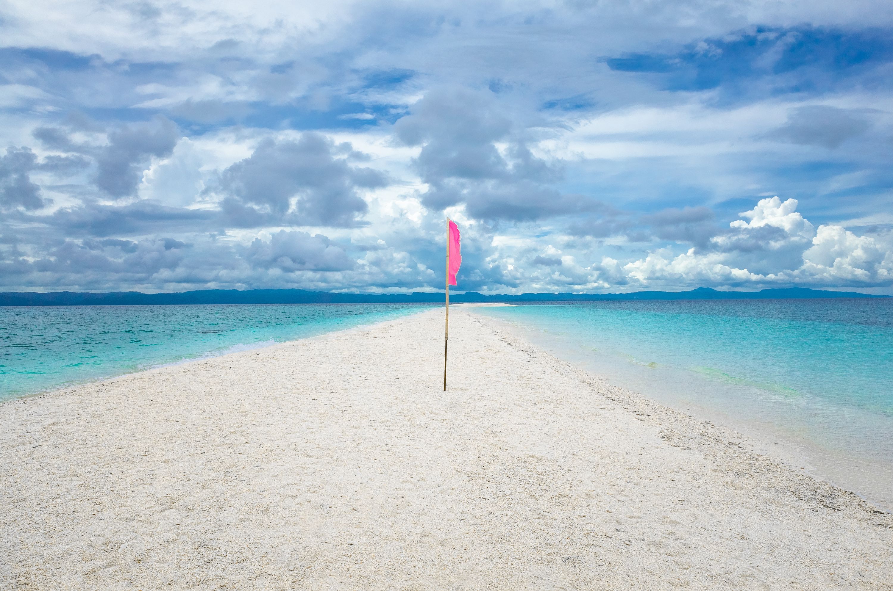 Une plage avec un drapeau rose, une eau turquoise cristalline et des montagnes lointaines sous un ciel nuageux.