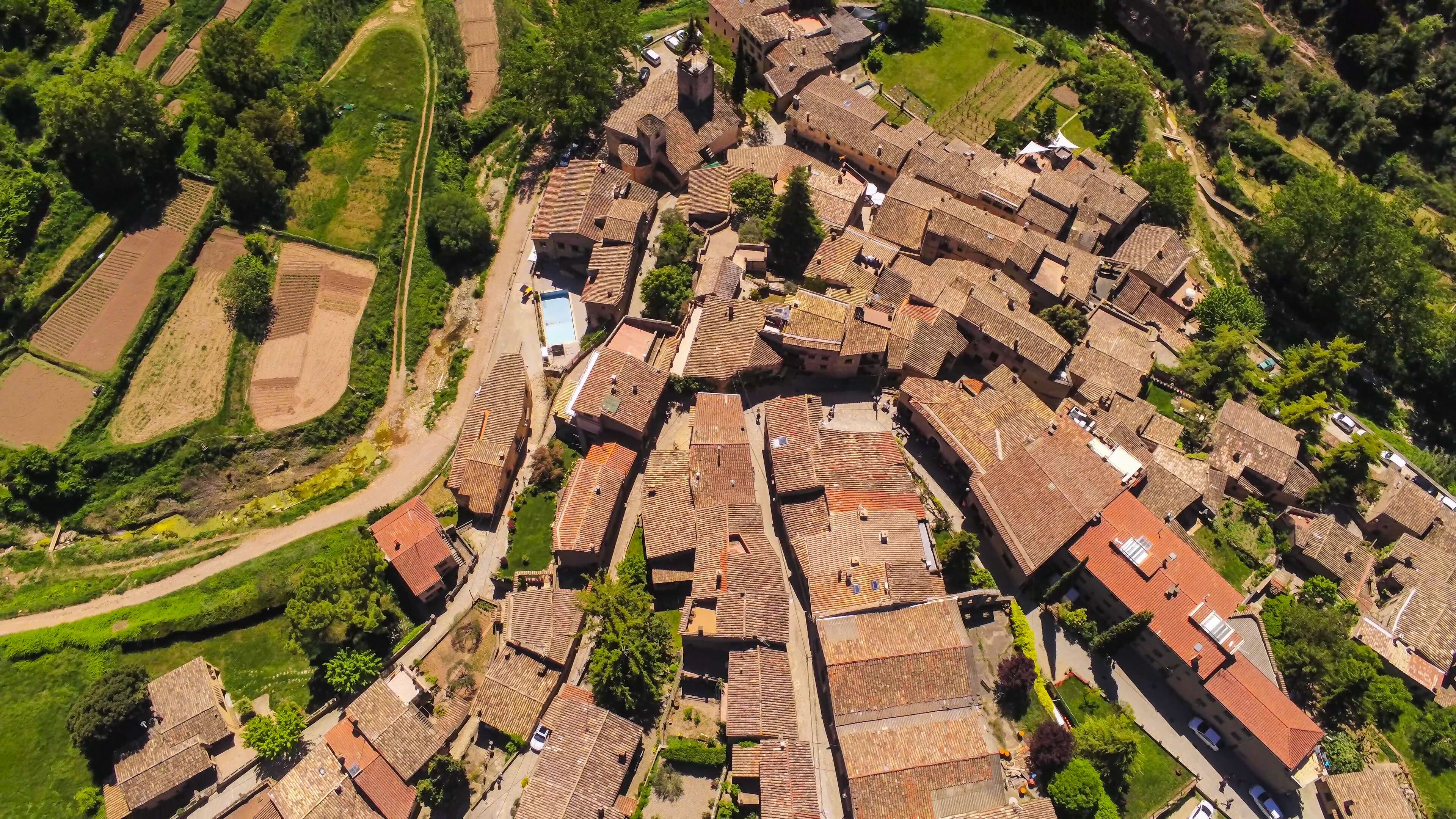 An aerial view of a historic village with terracotta-roofed buildings, narrow streets, and surrounding agricultural fields.