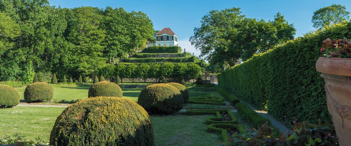 A landscaped garden with neatly trimmed hedges, spherical topiary, and a building with a red roof.