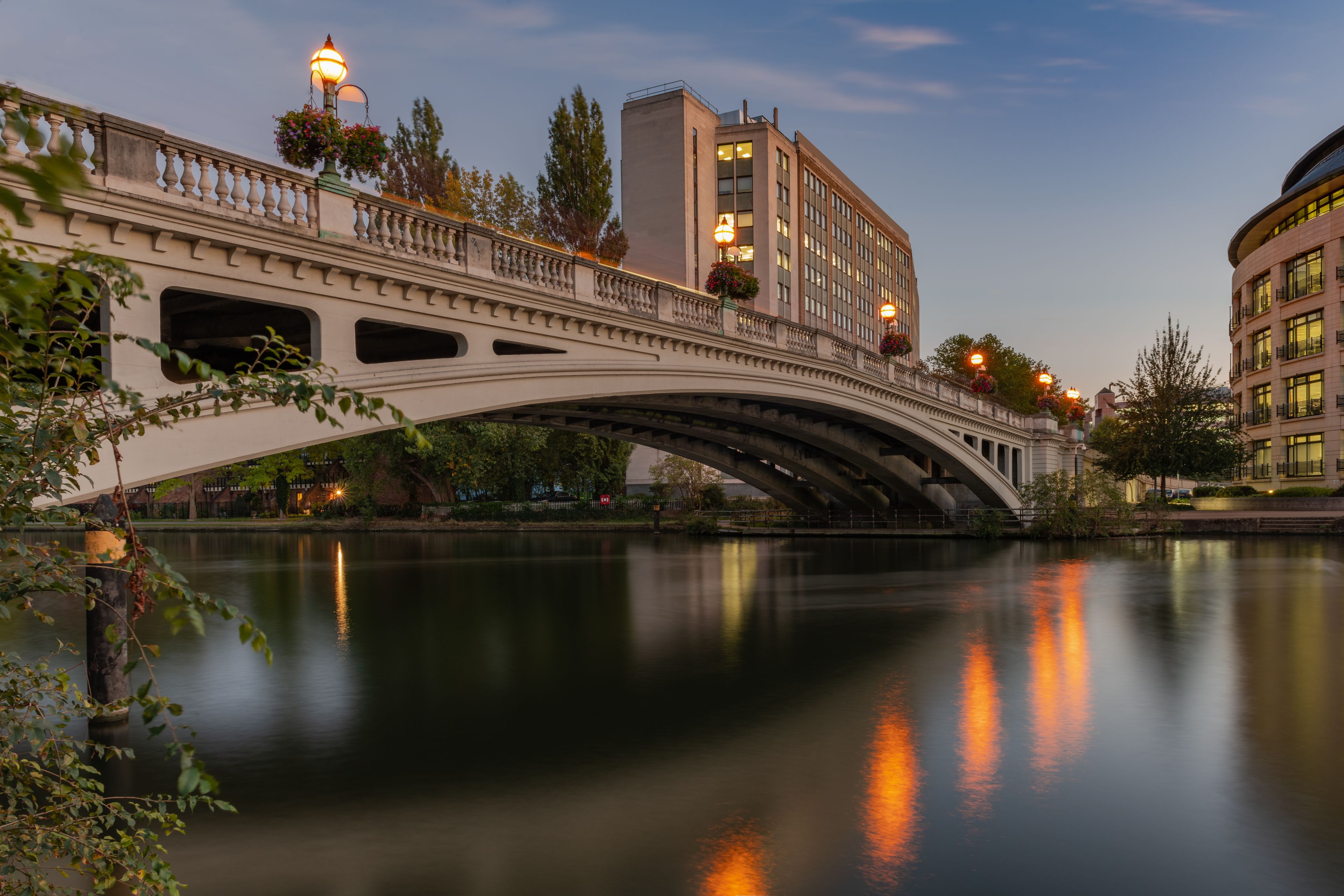 A bridge with ornate railings and flower baskets, spanning over a calm river with reflections of lights.