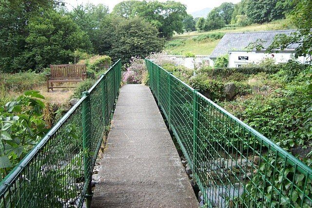 A narrow footbridge with green railings, surrounded by lush greenery and a white building in the background.