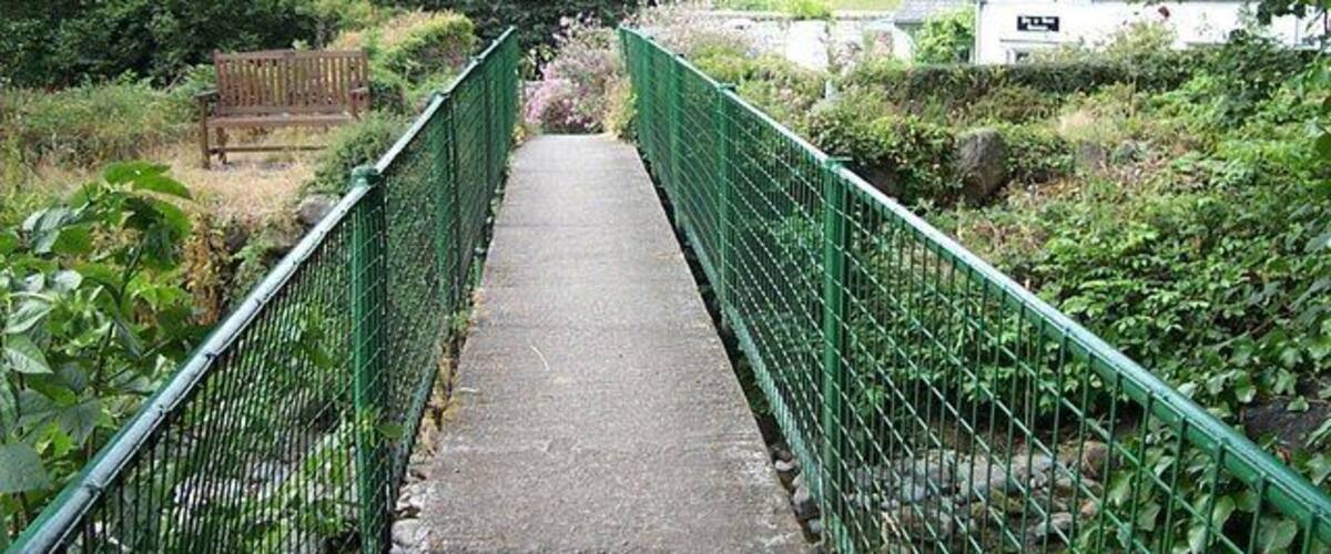 A narrow footbridge with green railings, surrounded by lush greenery and a white building in the background.