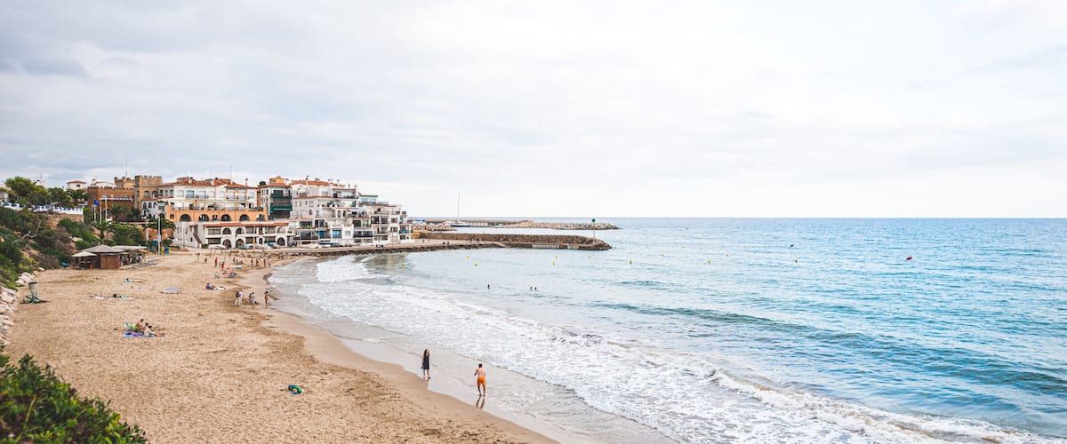 A beach with people walking and sunbathing, a pier extending into the water, and buildings in the background.