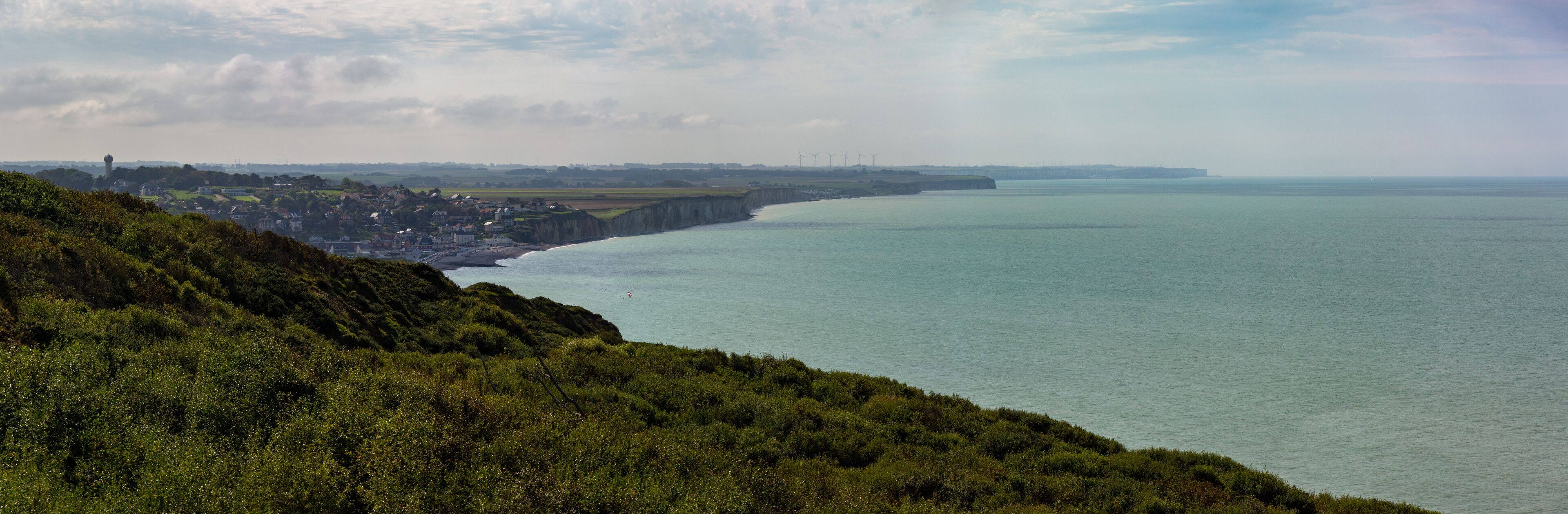 A coastal landscape with a cliff, a town, and wind turbines in the distance.