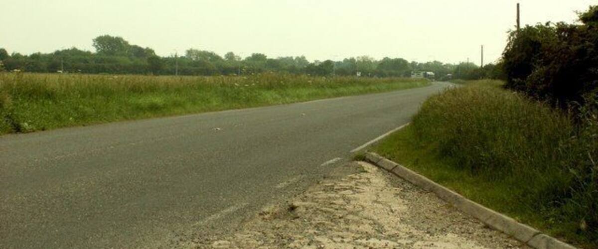 A rural road with grassy fields and a dirt shoulder.