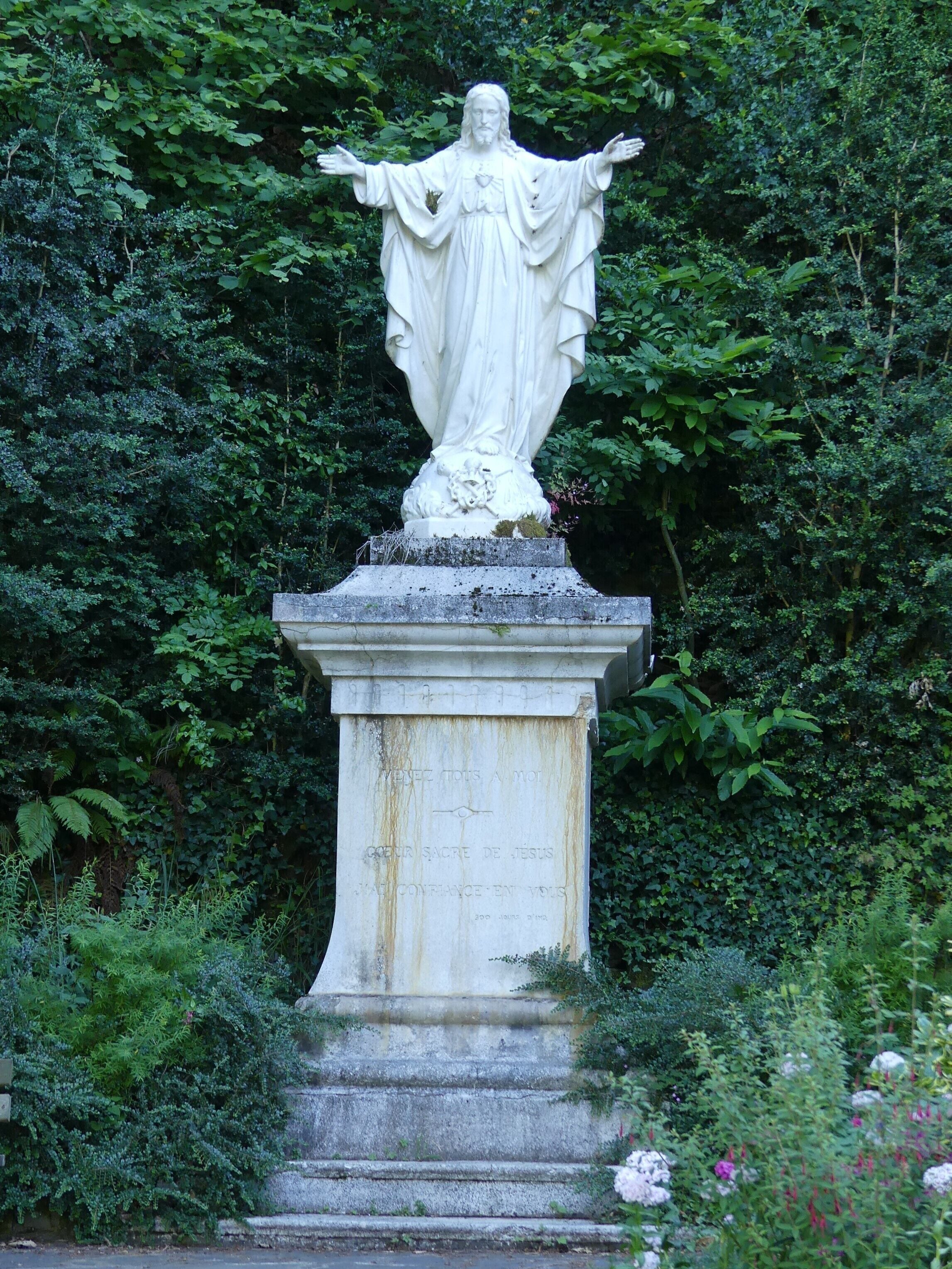 A white statue of a robed figure with outstretched arms, set on a pedestal with inscriptions, surrounded by lush greenery.