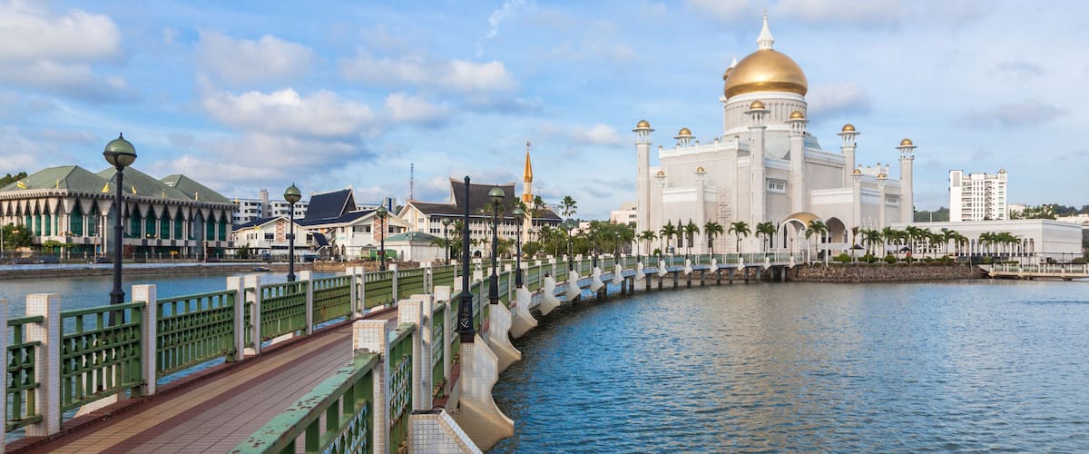 A waterfront promenade with a mosque featuring a golden dome, palm trees, and a bridge with green railings.