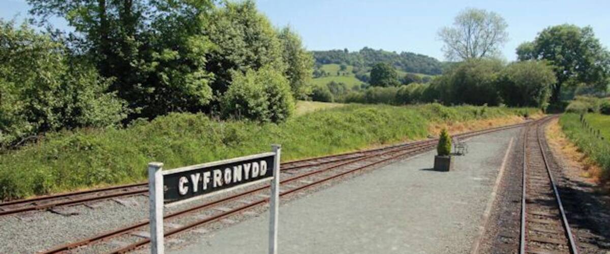 A railway track with a sign reading "CYFRONYDD" and a small potted plant on the platform.