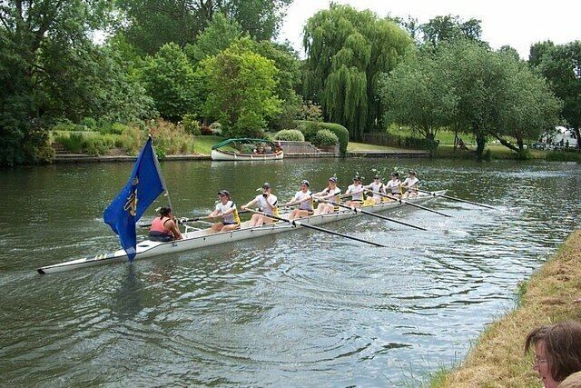 Cambridge - May Bumps, near to Fen Ditton, Cambridgeshire, Great Britain. the white team celebrate their victory with flag flying and oars woman 2nd from left has celebratory drink.