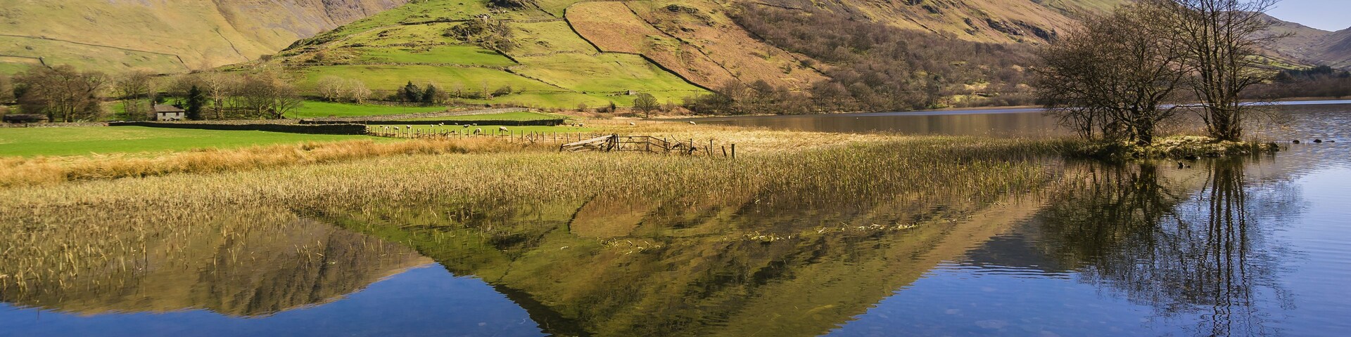 A calm lake reflecting a mountain range, a clear sky, and a grassy landscape with a small building.