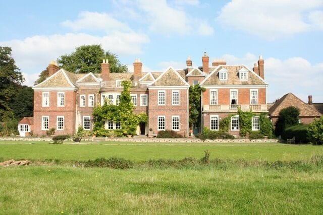 A large brick manor with multiple chimneys, surrounded by a green field and trees.