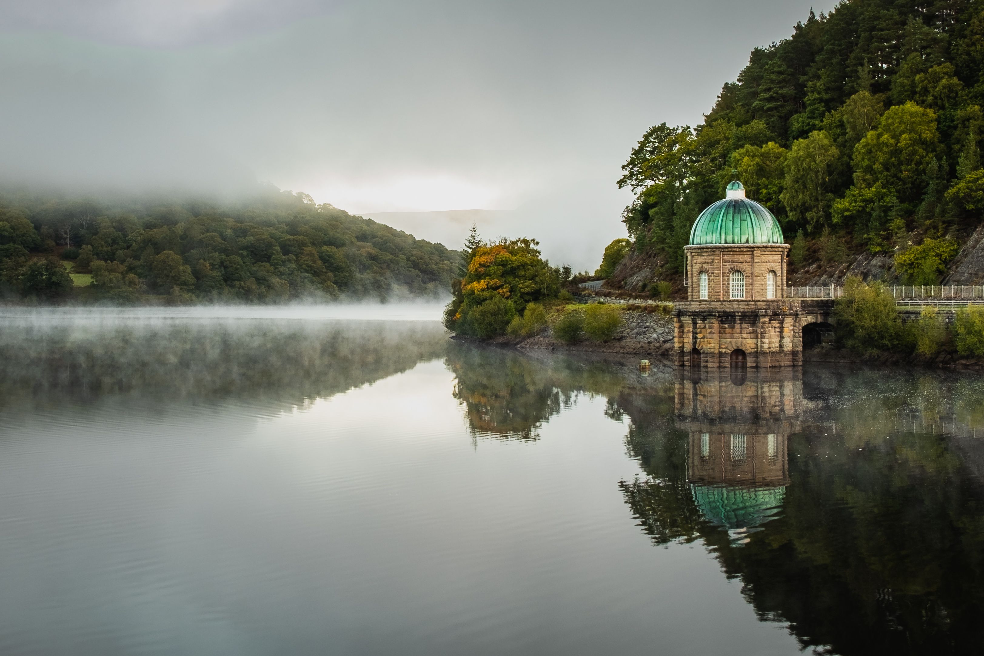 Un lac paisible, avec un petit bâtiment surmonté d’un dôme vert, entouré d’une végétation luxuriante et de collines.