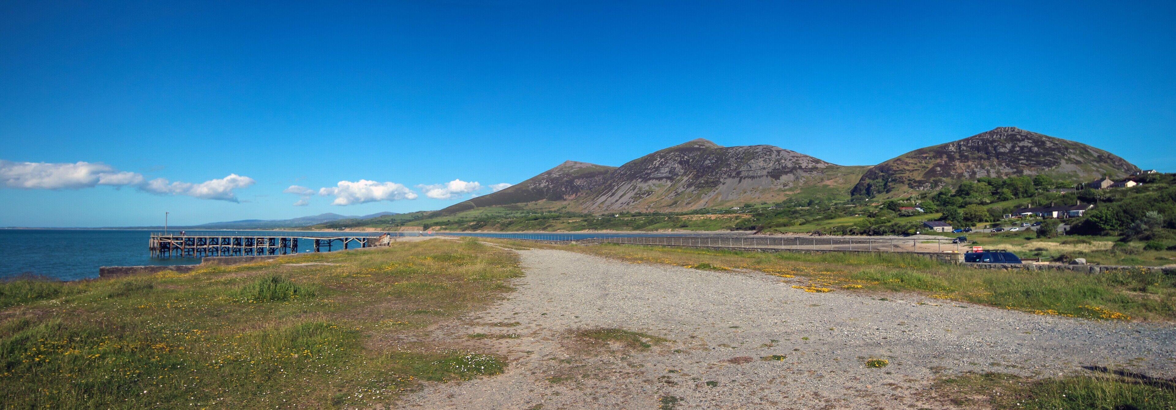 A coastal path leading to a pier, with mountains and houses in the background.