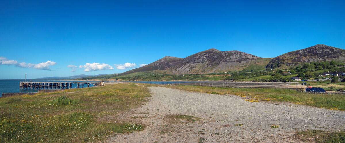 A coastal path leading to a pier, with mountains and houses in the background.