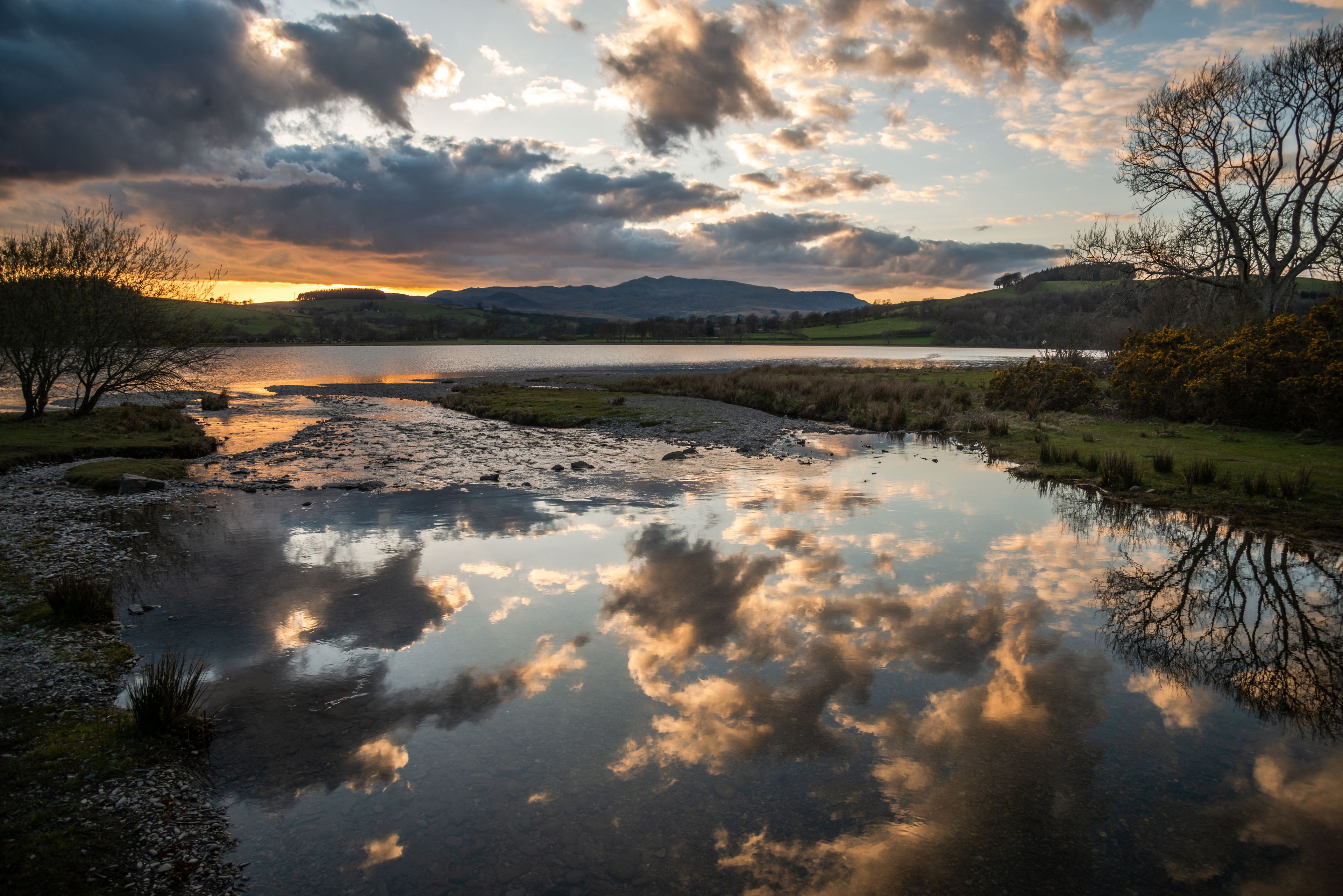 Un río tranquilo que refleja nubes, rodeado de colinas y escasos árboles.