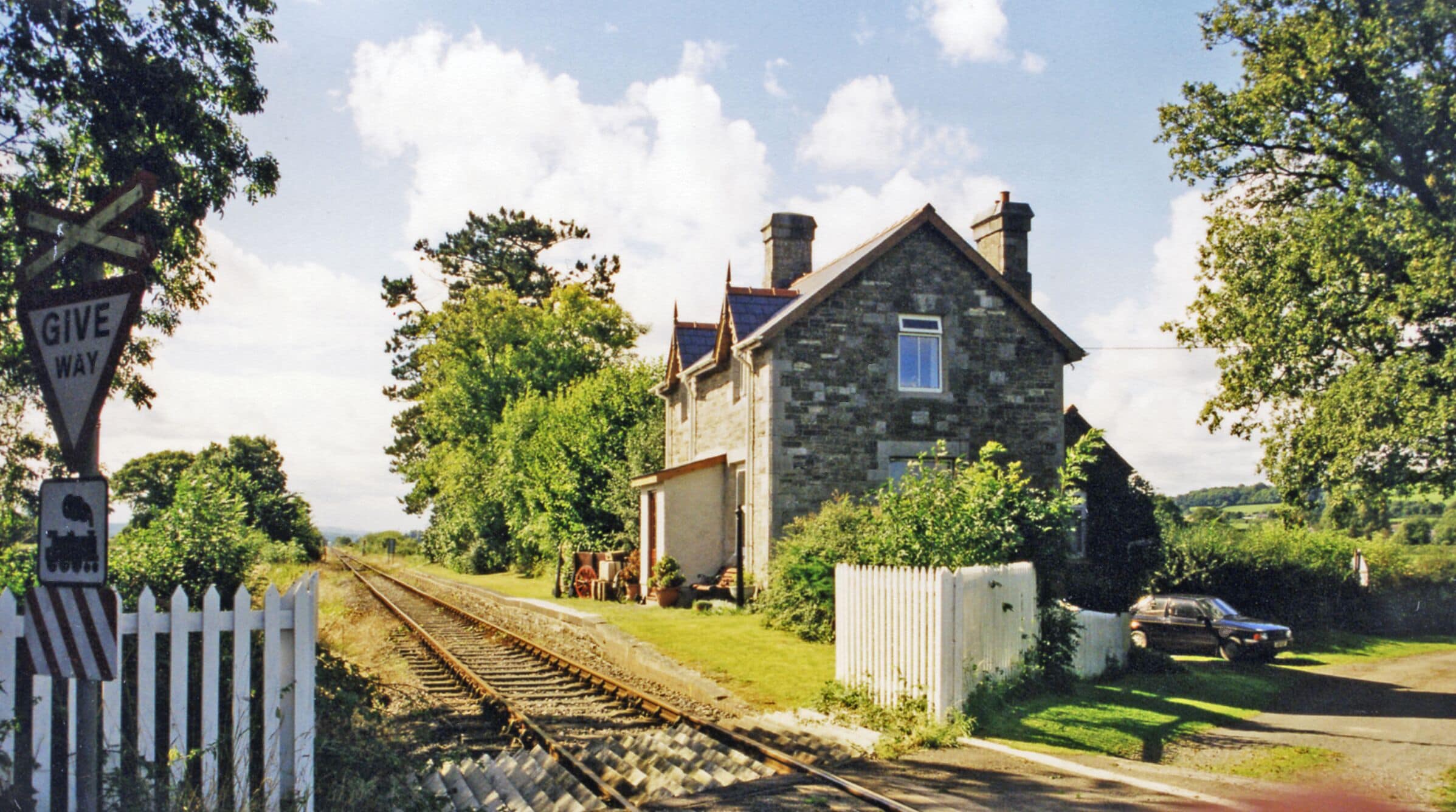 A stone house with a white picket fence, a railway crossing, and a car parked on the road.
