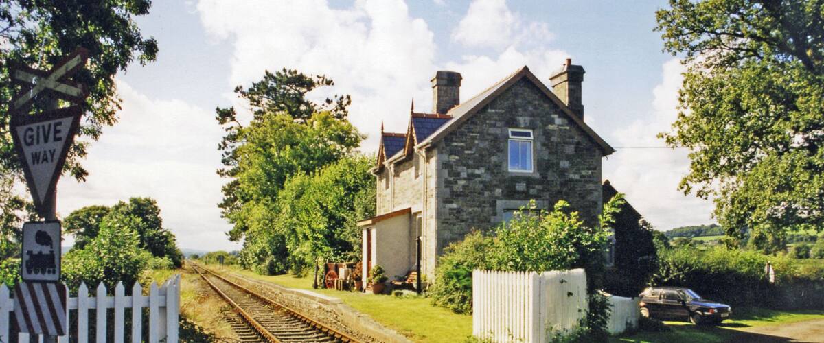 A stone house with a white picket fence, a railway crossing, and a car parked on the road.