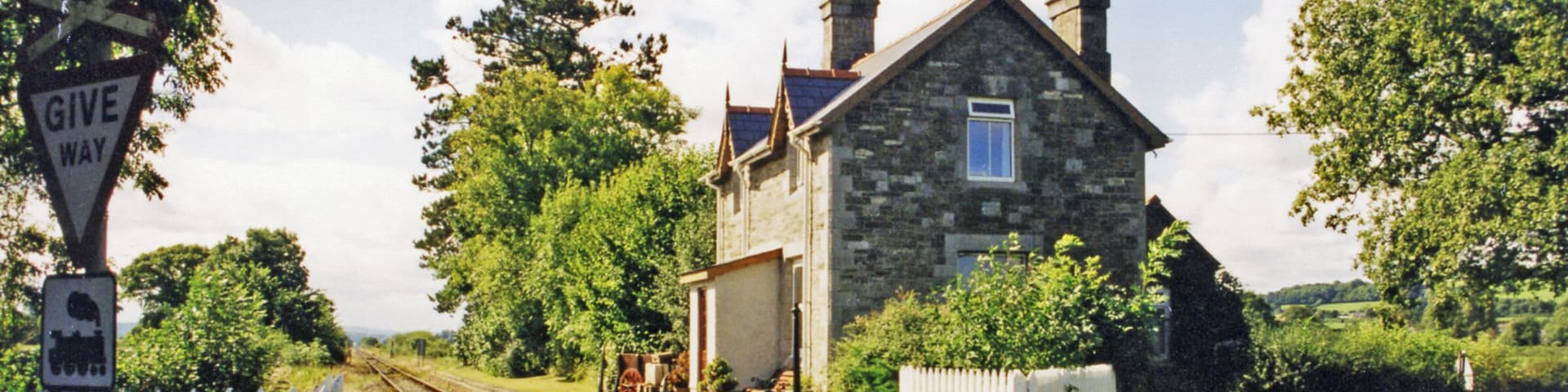 A stone house with a white picket fence, a railway crossing, and a car parked on the road.