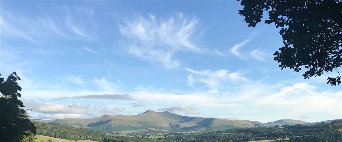 A rural landscape with rolling hills, scattered trees, and a distant mountain range under a partly cloudy sky.