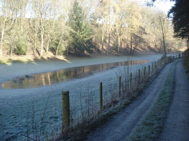 Un sendero cubierto de nieve junto a un río tranquilo, rodeado de árboles y una valla de madera.