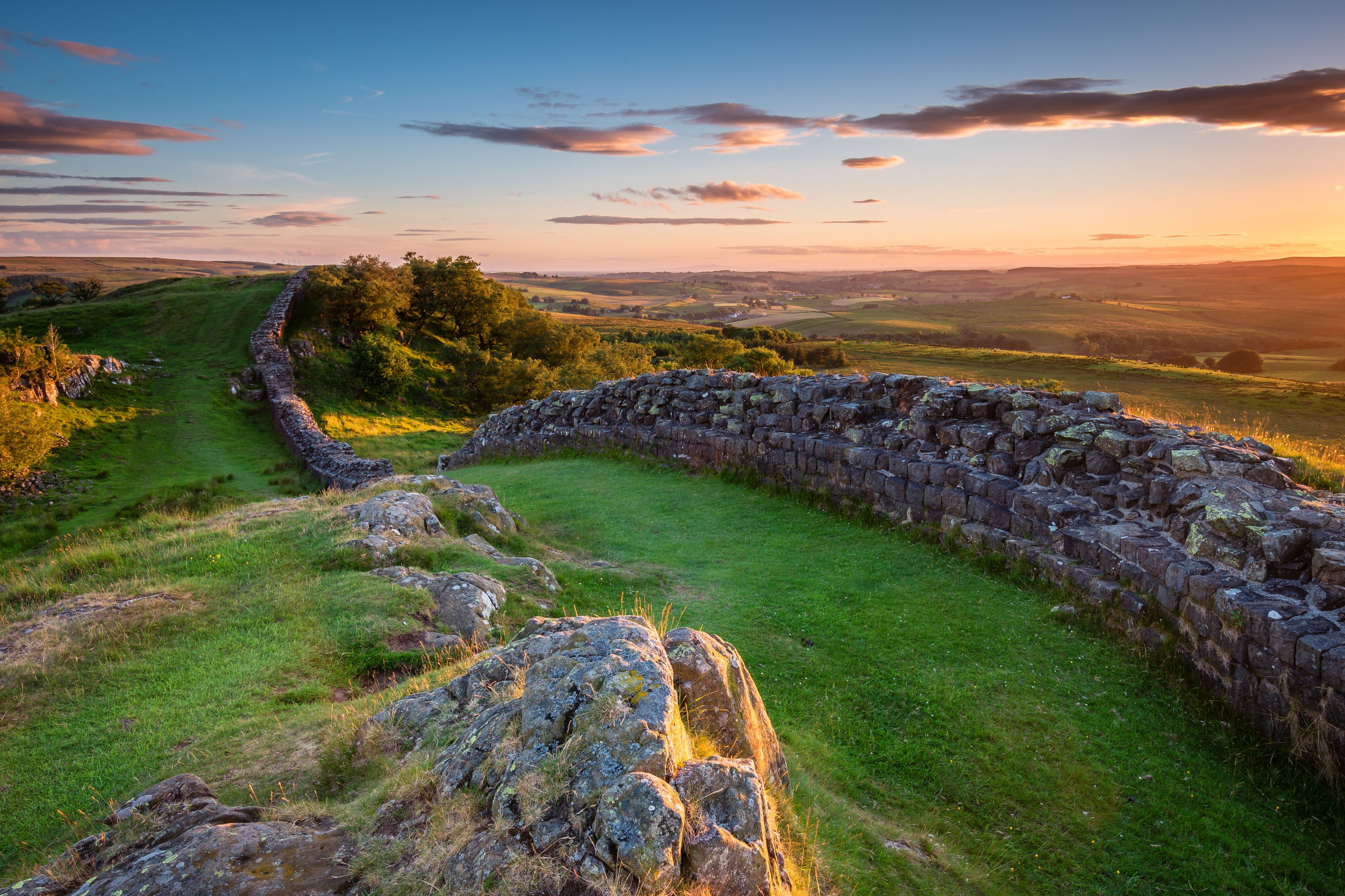 A stone wall curving through a grassy landscape with trees and a sunset in the background.