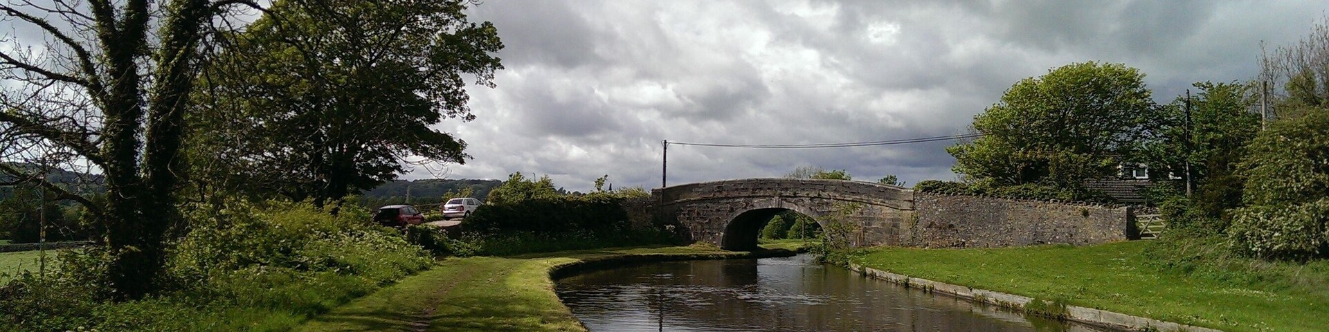 A narrow canal with a stone bridge, lush greenery, and a cloudy sky.
