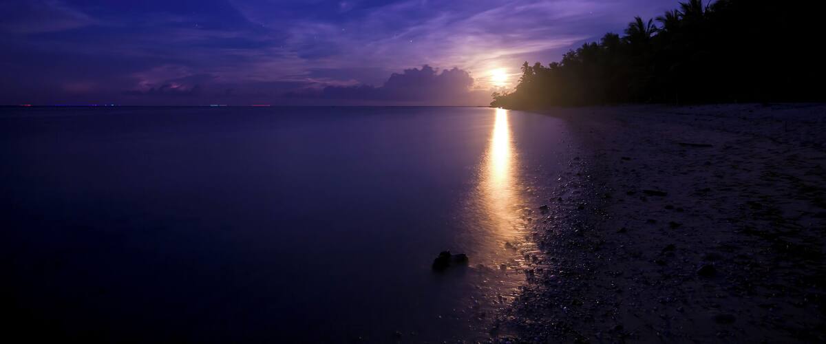 A beach at sunset with a calm sea, a visible boat, and a distant city skyline.