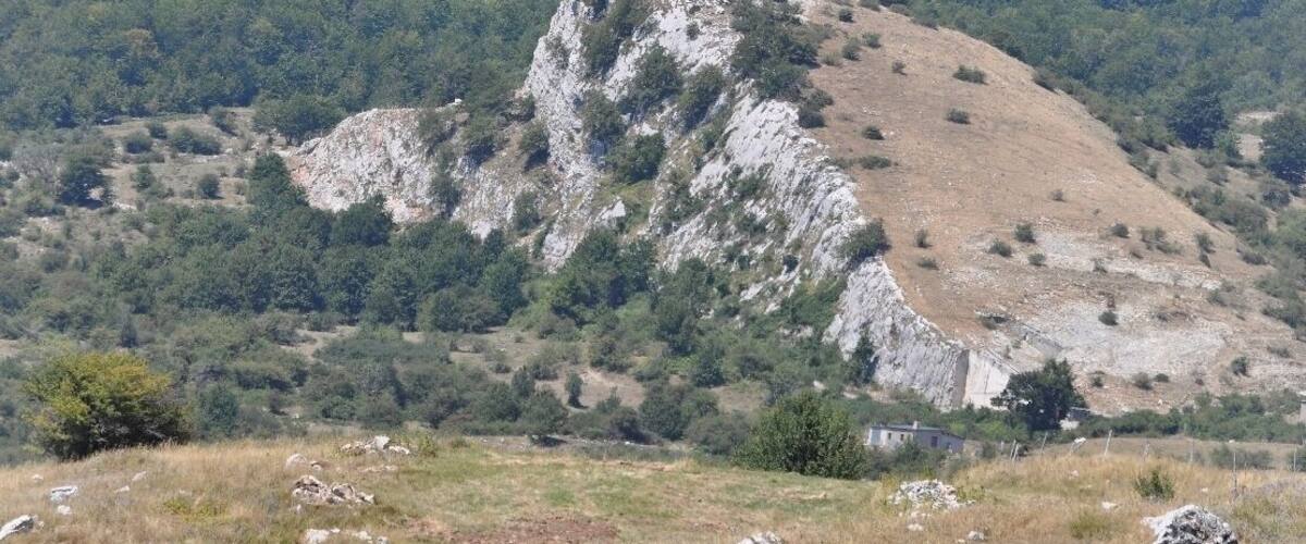 A rugged mountain with a prominent rocky peak, surrounded by dense forest and a grassy valley.