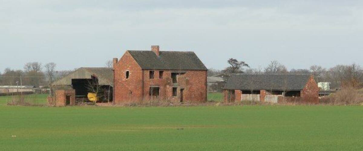 An abandoned brick house with a chimney, surrounded by open fields and distant trees.