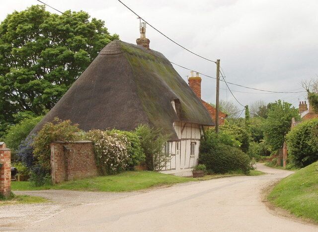 A thatched-roof house with a white exterior, a narrow road, and lush greenery in the background.