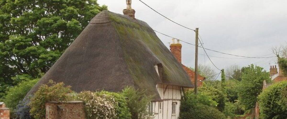 A thatched-roof house with a white exterior, a narrow road, and lush greenery in the background.