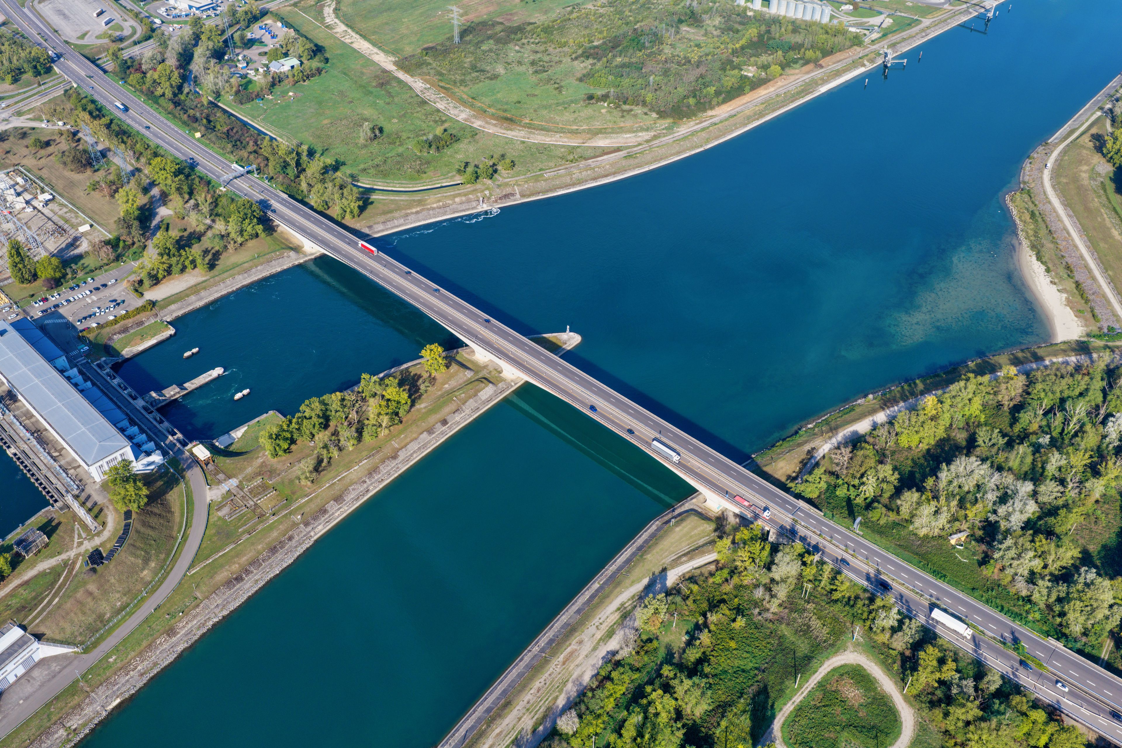 Luchtbeeld van een brug over een rivier, met groene oevers en industriële gebouwen.