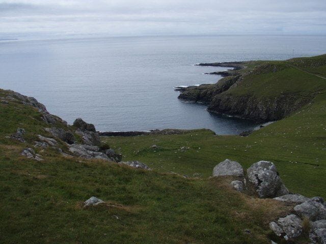 Paisaje costero con colinas cubiertas de hierba, afloramientos rocosos y mar en calma.