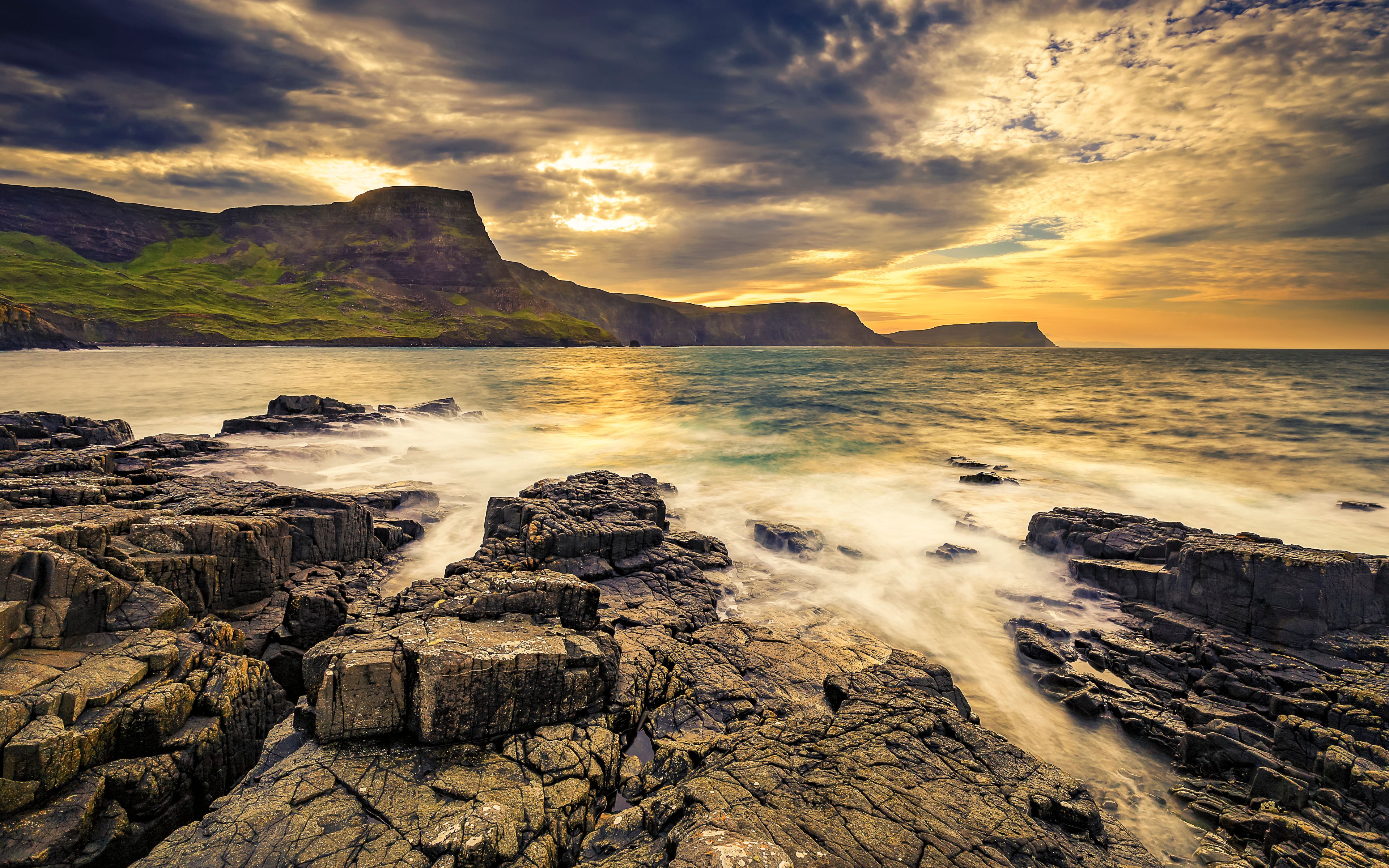 A rocky coastline with waves crashing against the rocks, a cliff in the background, and a sunset sky.