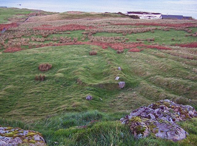 Ruin below Dun Chaich There is much evidence of lazybeds and ruined walls and buildings in this area. Here, looking down from the scant remains of Dun Chaich, the outline of a building and wall shows as humps in the grass. They may well have been built using stone robbed from the dun.