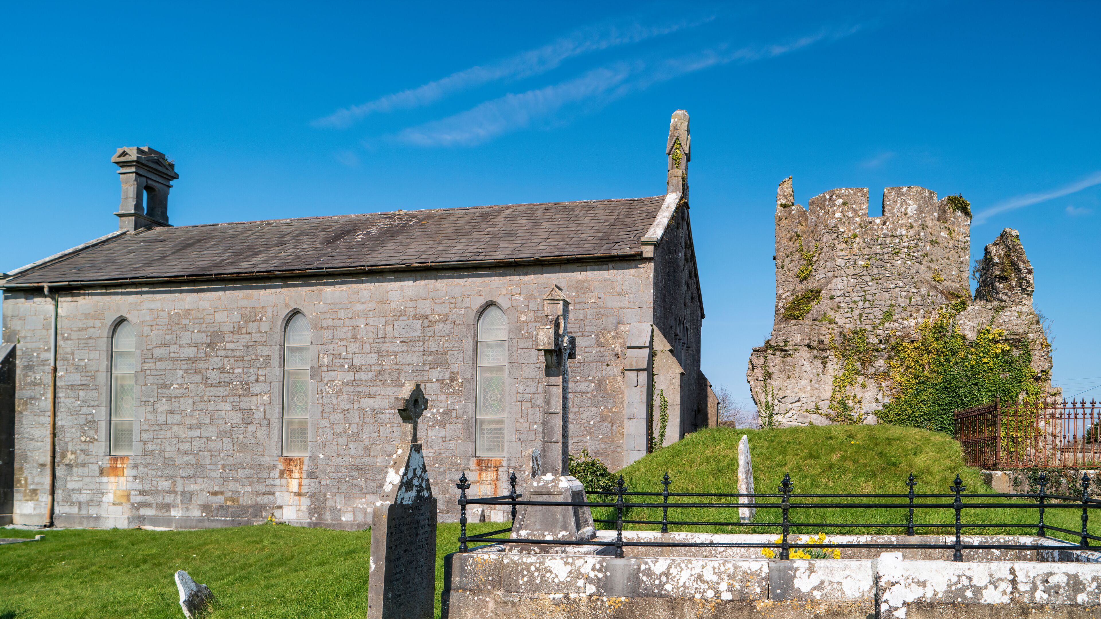 A historic stone church with arched windows and a bell tower, adjacent to a ruined castle wall with ivy.